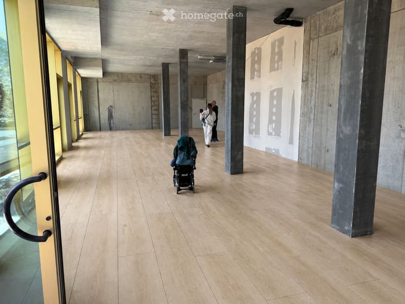 Modern hallway with light wooden flooring, concrete pillars, and large glass windows on one side. A stroller is visible in the foreground, and a few people are walking in the background.