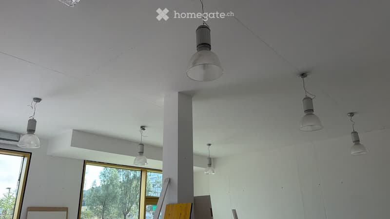 Empty room with white walls, exposed ceiling beams, and several hanging pendant lights. Large windows provide natural light, and the space appears to be a commercial or gallery area.