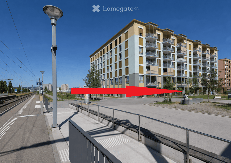 Modern multi-story residential building with balconies and large windows, located near a railway track and street with street lamps.