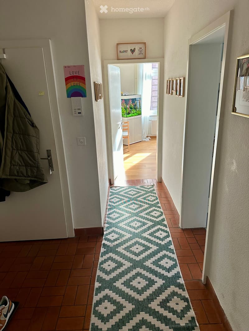 Hallway with tiled floor, patterned runner rug, open door leading to another room, coat hanging on the left, and framed pictures on the right wall.