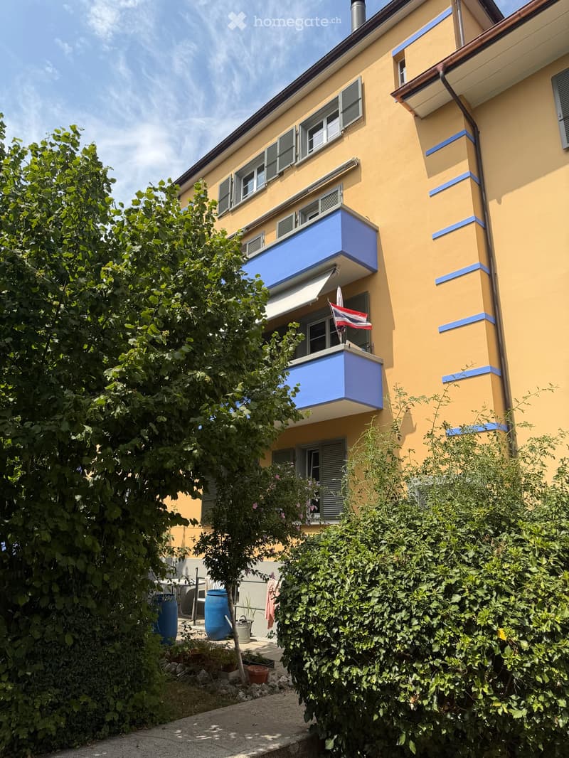A yellow multi-story building with blue balconies and shutters, surrounded by greenery and outdoor furniture.