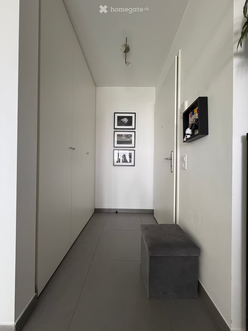 Modern hallway with white walls, grey tiled floor, and a small bench. Features framed pictures on the wall and a wall-mounted light fixture.