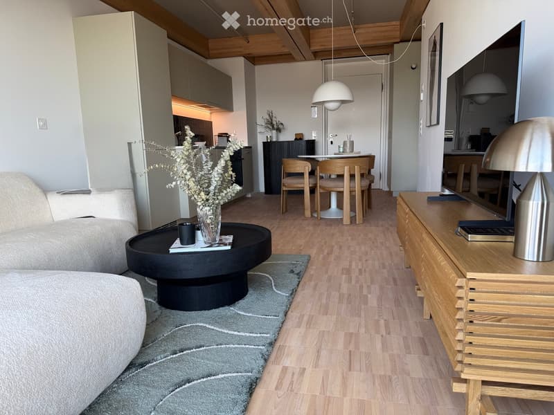 Modern living room with a grey sofa, coffee table, dining area, and kitchen in the background. Features wooden flooring, pendant lights, and a sleek TV stand.
