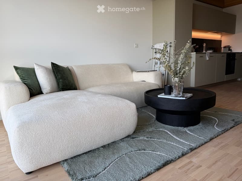 Modern living room with a beige sectional sofa, green throw pillows, a round black coffee table, a vase with dried flowers, and a small kitchen area with grey cabinets.