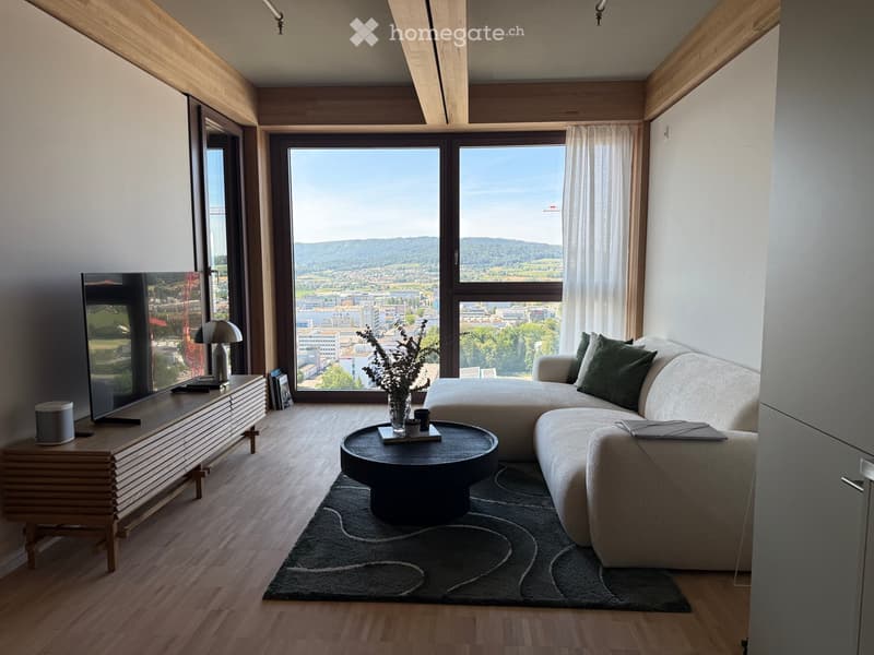 Modern living room with large windows offering a scenic view, featuring a grey sectional sofa, a round coffee table, a side table with a lamp, and a television on a wooden stand.
