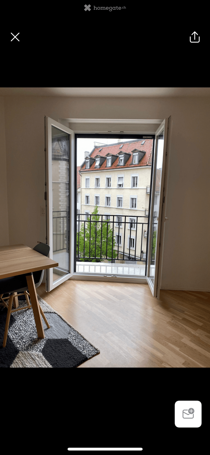 Bright room with wooden flooring, a small table and chair, and a rug near the door leading to a balcony with a view of a building.
