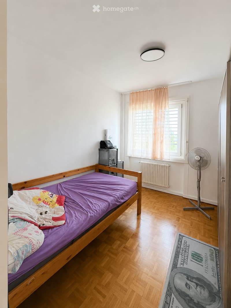A small bedroom with a wooden bed, purple bedding, a standing fan, a radiator, and a laptop on a small table. The room has wooden flooring and a window with light-colored curtains.