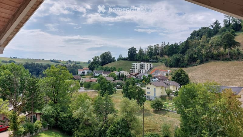 View from a balcony or terrace overlooking a residential area with houses, trees, and open fields under a partly cloudy sky.