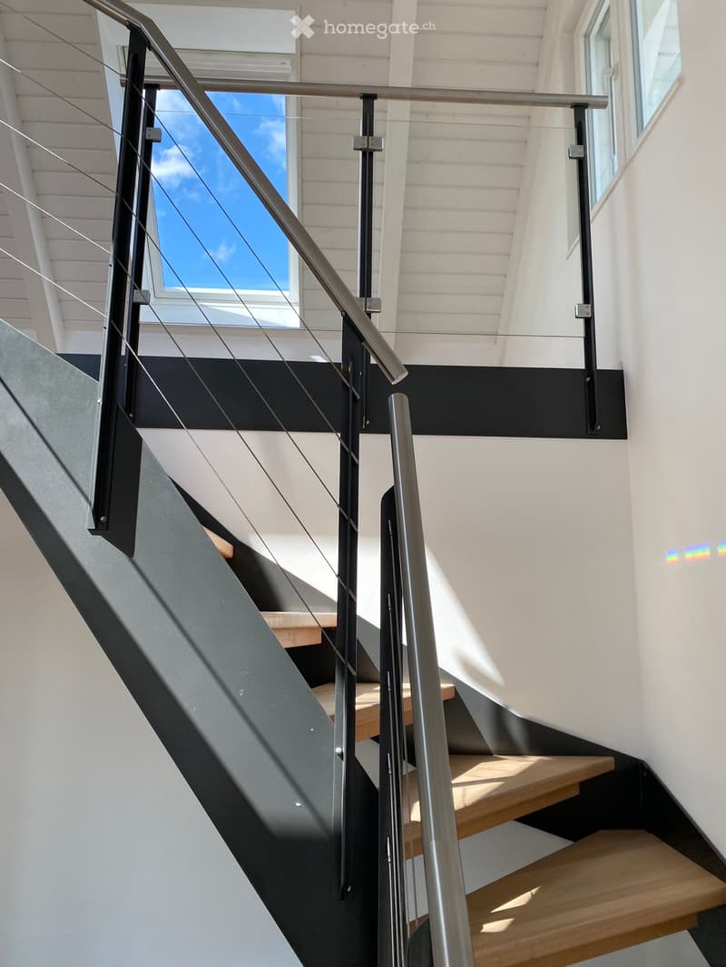 Modern staircase with wooden steps, metal railing, and glass panel, set against white walls and a slanted ceiling with wooden paneling.