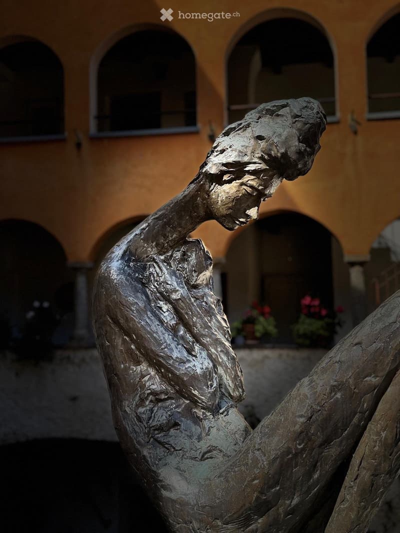 Bronze sculpture of a human figure in a dynamic pose, set against a backdrop of a historic building with arched windows and potted plants.