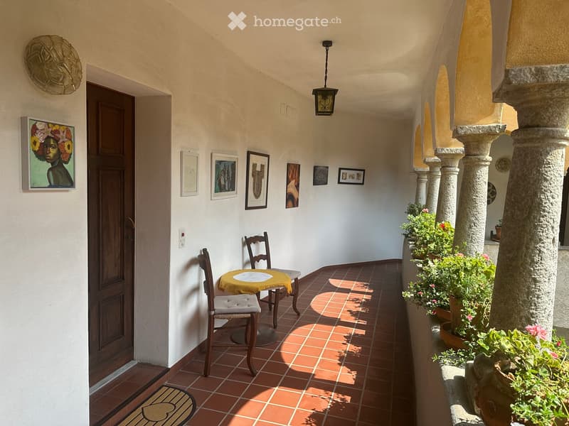 Balcony with terracotta tiles, a small round table with a yellow tablecloth, two wooden chairs, and several potted plants along the railing.
