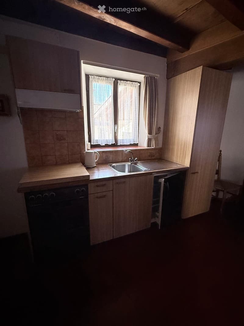 Compact kitchen with wooden cabinets, a sink, an oven, and a refrigerator. Light-colored countertops and a window with curtains provide natural light.