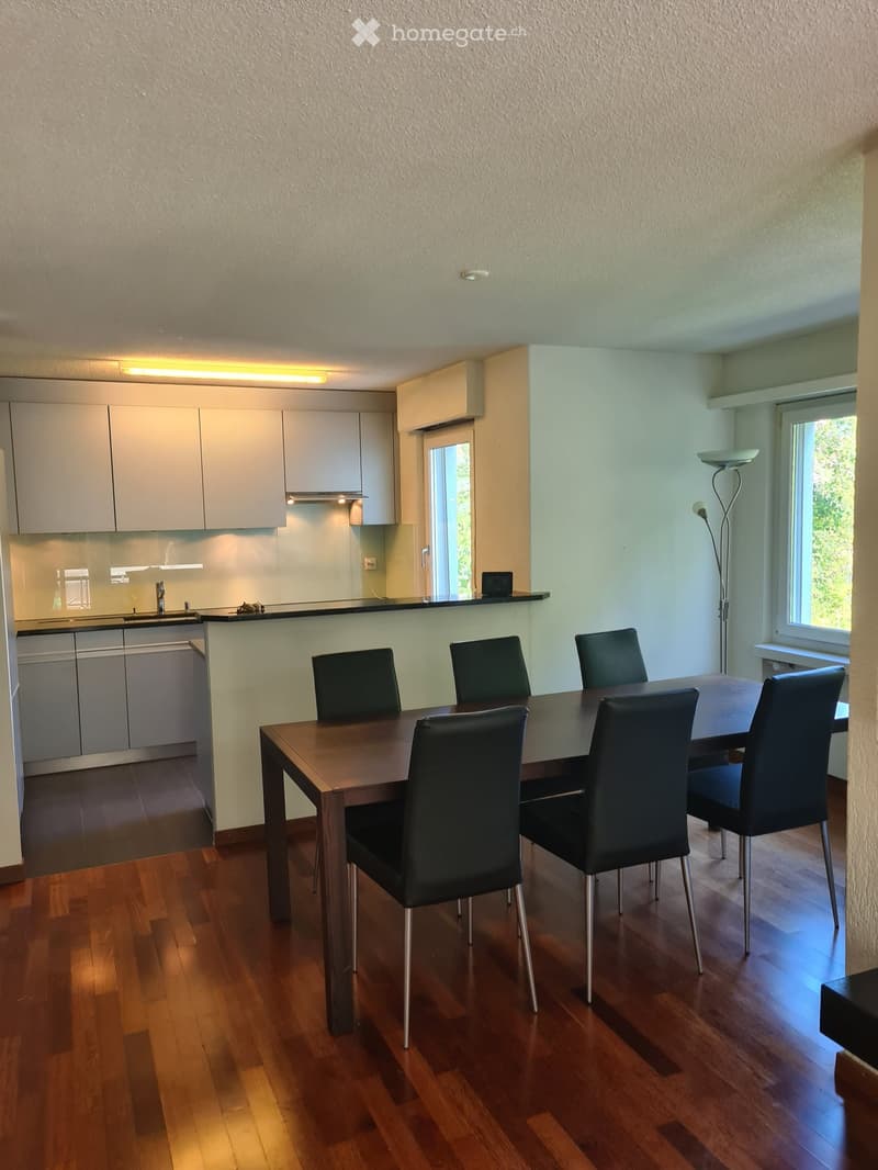 Modern dining area with a wooden dining table and six black chairs, adjacent to a sleek kitchen with white cabinets and stainless steel appliances.