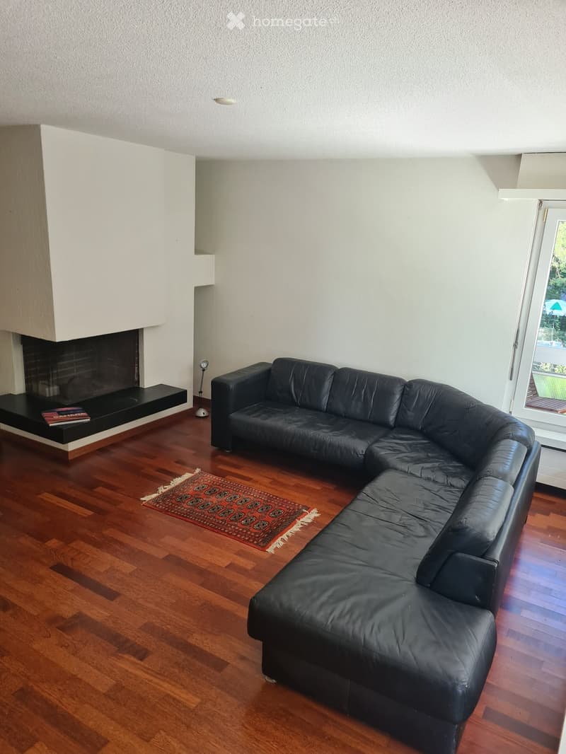 Modern living room with a black leather sectional sofa, a fireplace with a black mantel, a small wooden table, and a patterned rug on a wooden floor.