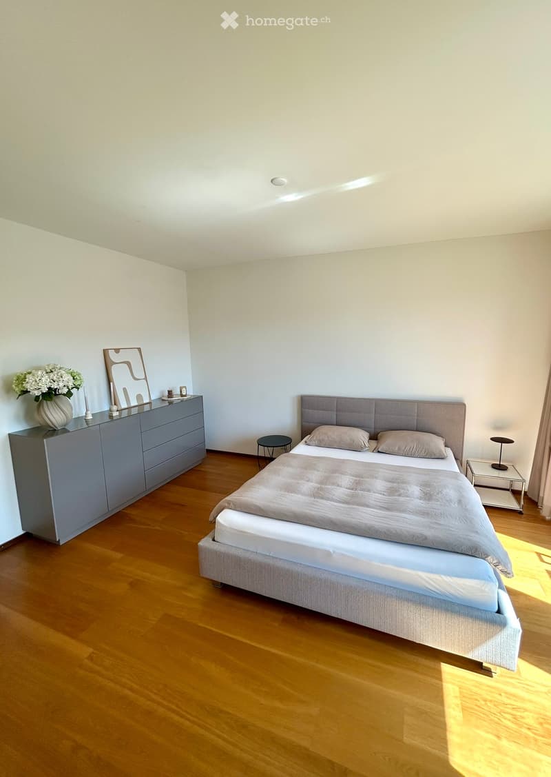 Modern bedroom with wooden flooring, a grey upholstered bed, nightstand, mirror, and decorative vase on a dresser.