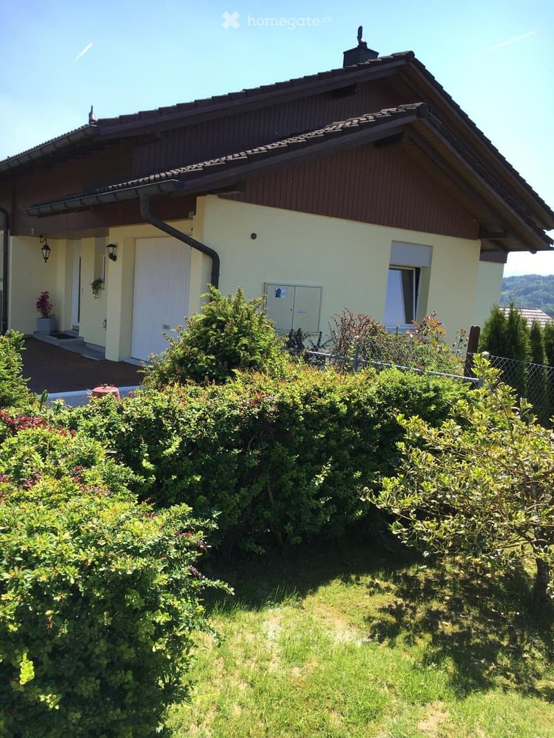 Two-story house with a sloped roof, light yellow walls, and a wooden facade on the upper level. The house features a small porch with a white door, a window with white shutters, and a small garden area with various plants and shrubs.