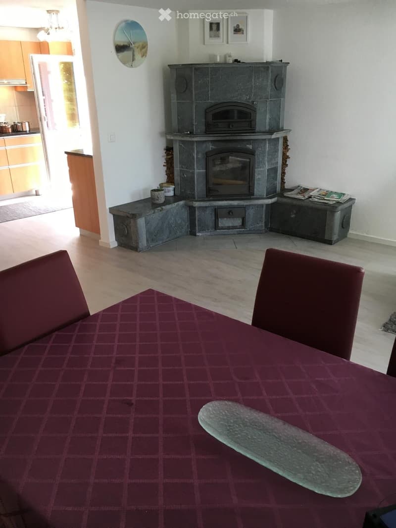 Dining area with a large table covered in a red checkered tablecloth, two red chairs, a stone fireplace with a built-in bench, and a small kitchen area visible in the background.