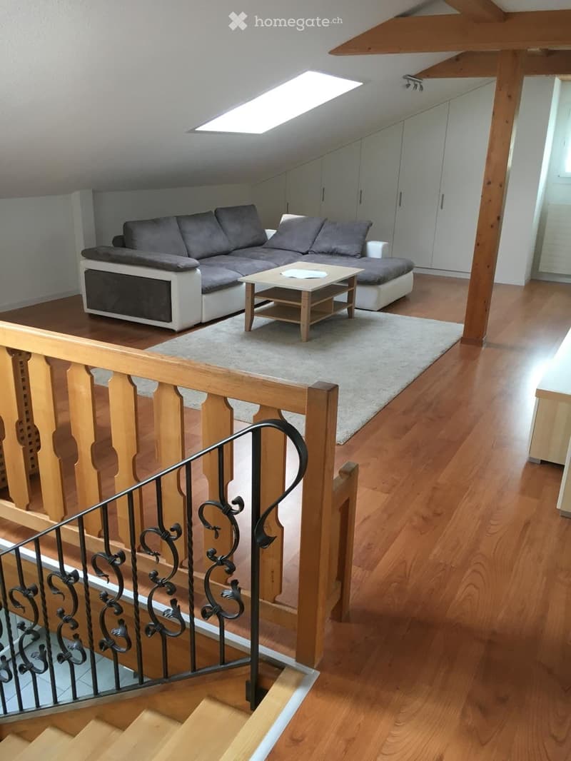 Modern living room with grey sectional sofa, white and grey accents, wooden coffee table, and area rug. Wooden staircase with metal railing leads to another level.