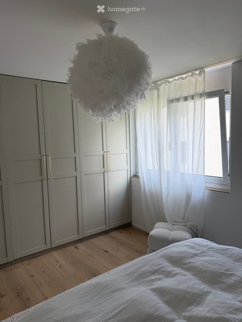 A modern bedroom with a large bed, a wardrobe, a chair, and a decorative light fixture. The room features wooden flooring and large windows with white curtains.