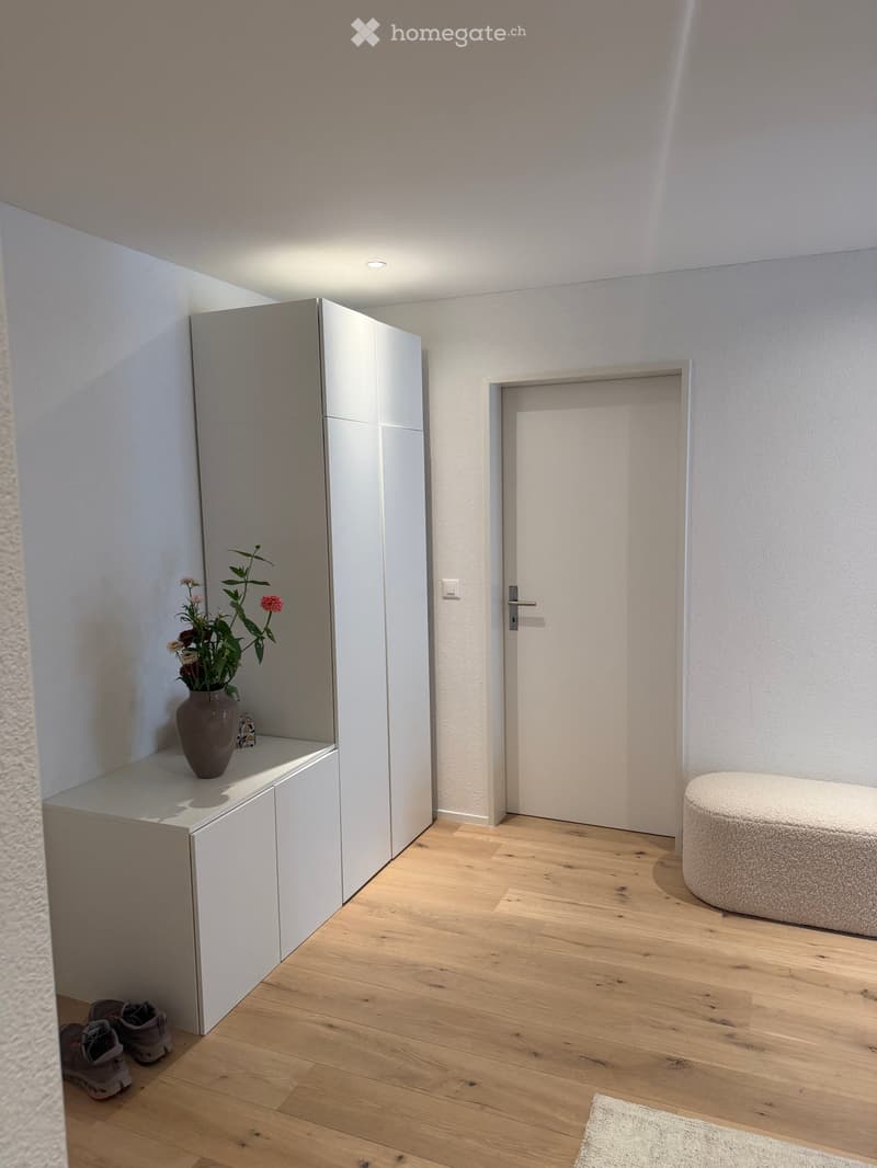 A modern, minimalist hallway with light wooden flooring, white walls, a built-in shoe rack, a small round stool, and a vase with flowers on the shelf.