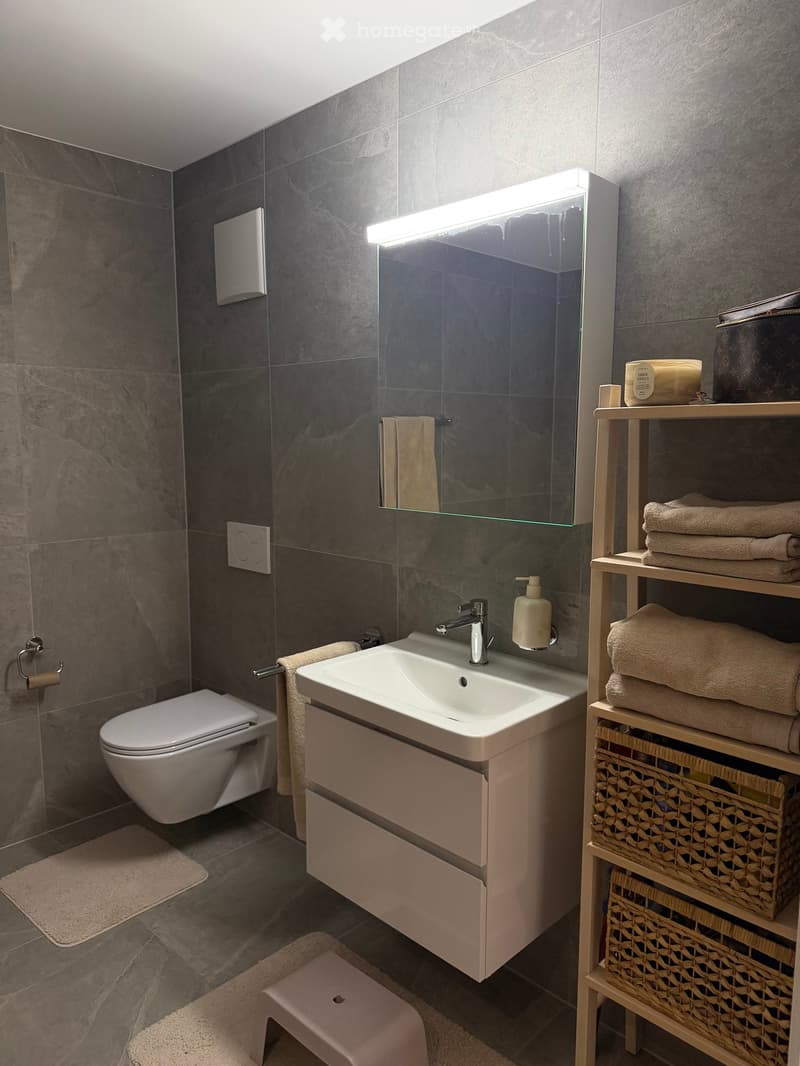Modern bathroom with grey tiled walls, a white sink with a chrome faucet, a large mirror with built-in lighting, a toilet, a wooden shelf with towels and a woven basket, and a small pink stool.
