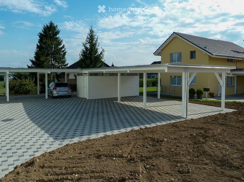 Two-story yellow house with a gray roof, white-framed windows, and a covered driveway with a parked car. The driveway is paved with interlocking bricks, and there is a landscaped garden area with small trees and shrubs.