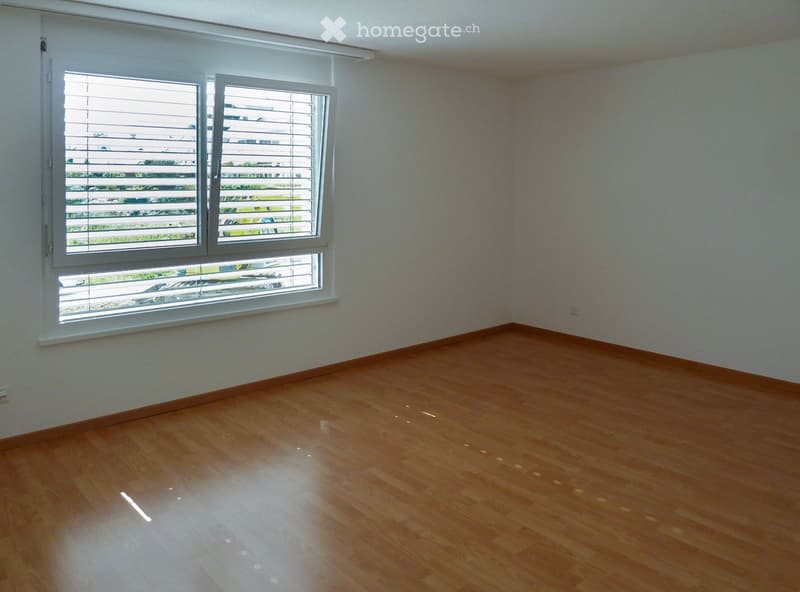 Empty room with wooden flooring and large windows with blinds, showing a view of the outside area.