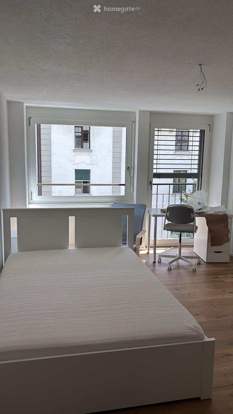Modern bedroom with a single bed, a small desk with a chair, and a cabinet. Large windows offer a view of the balcony and the street outside.