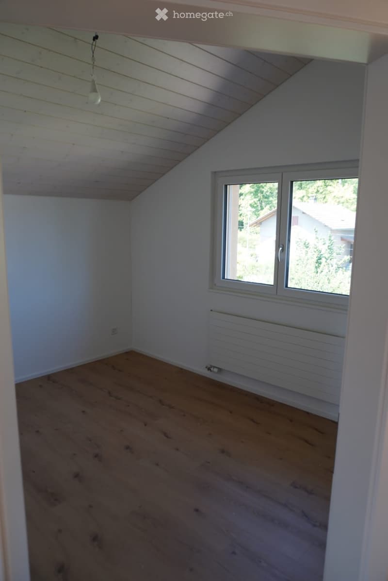 Empty attic room with wooden flooring, white walls, a single window, and a radiator. The ceiling has exposed wooden beams and a hanging light fixture.