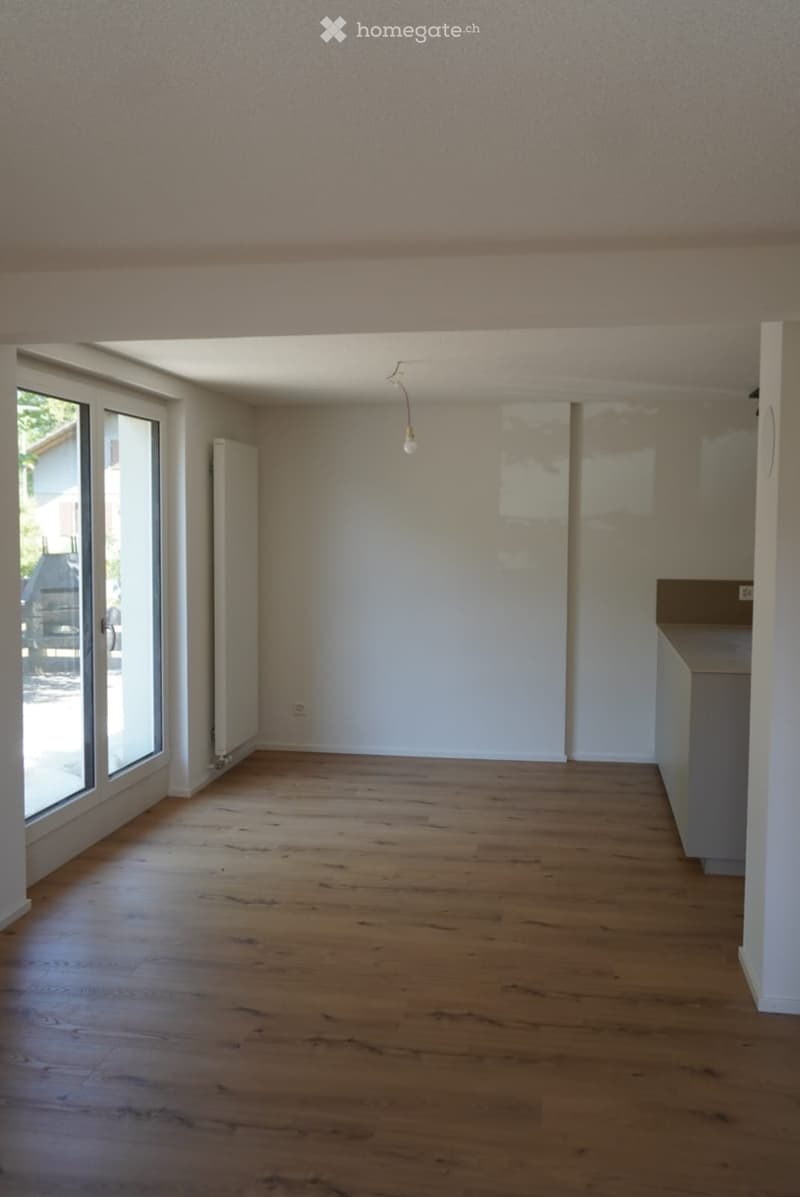 Empty room with wooden flooring, white walls, large windows, and a radiator visible on one wall.