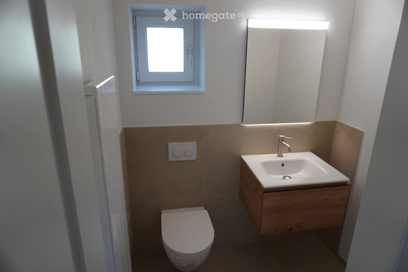 Modern bathroom with a toilet, sink, and mirror. The sink has a wooden cabinet underneath and a sleek chrome faucet. The walls are tiled in a light brown color, and there is a small window above the sink.