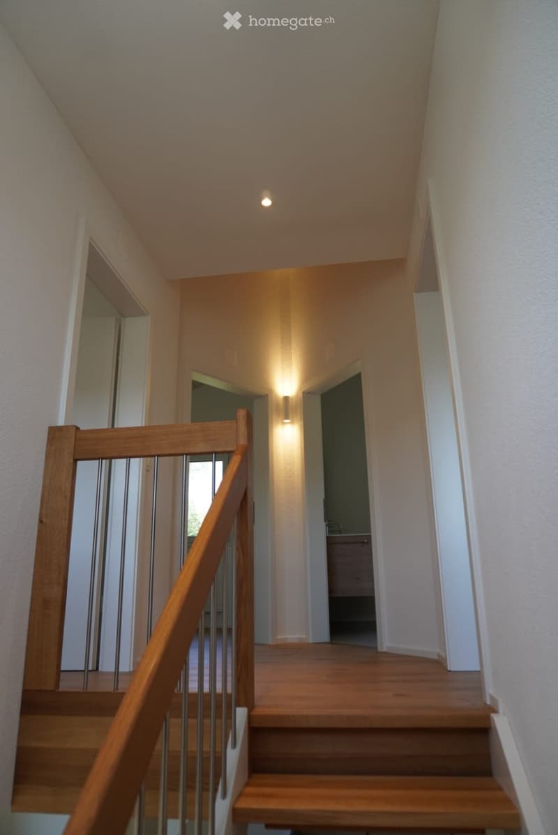 Wooden staircase with metal railing, beige walls, and recessed lighting. The hallway leads to an open area with visible furniture in the background.