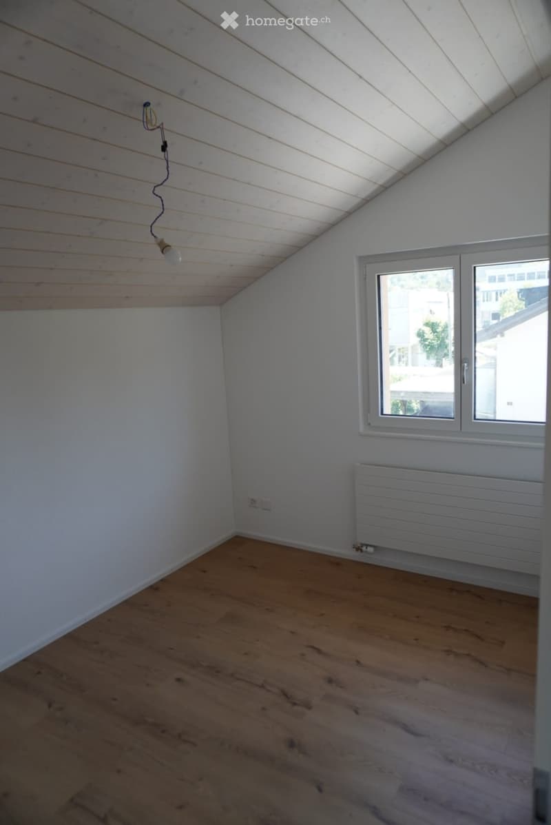 Empty attic room with wooden floor, white walls, slanted wooden ceiling, single window, and radiator.