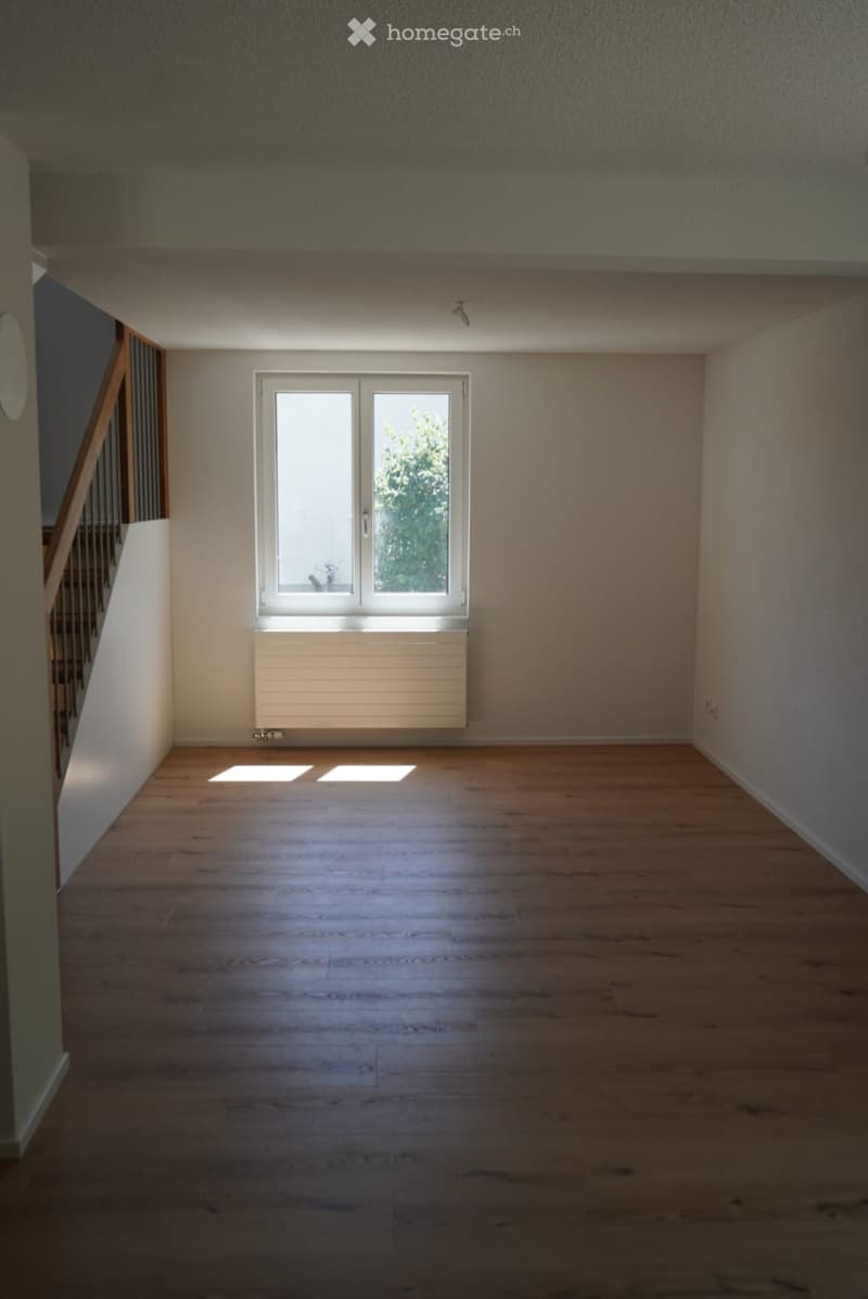Empty room with wooden flooring, white walls, and a large window with double doors leading outside. A radiator is visible below the window, and a staircase is partially visible on the left side.