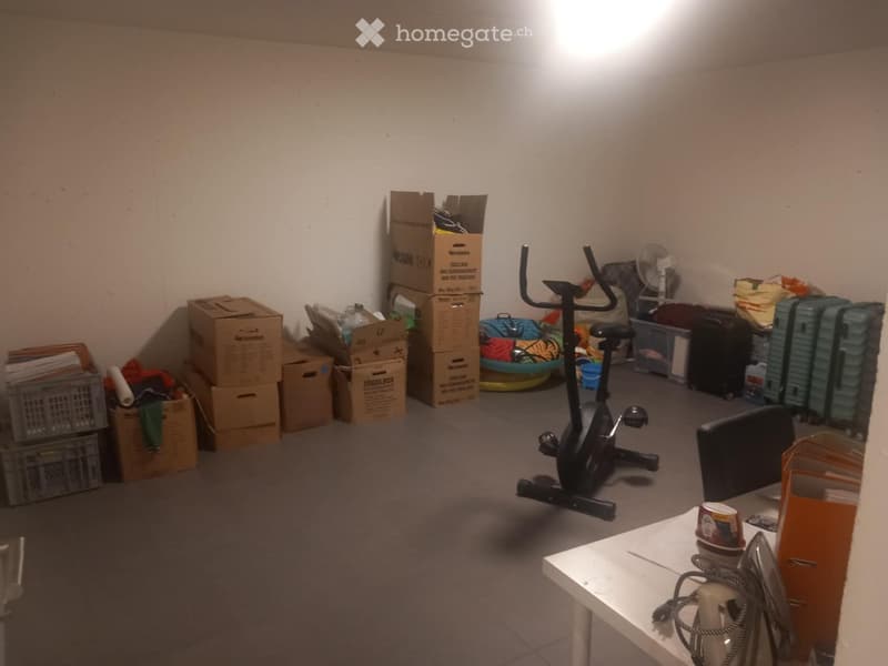 A room with an exercise bike, a table with a cup, a fan, several suitcases, and cardboard boxes stacked around.
