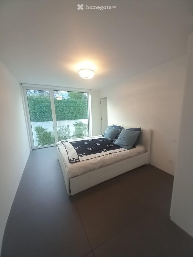 Modern bedroom with a white bed, blue pillows, and a dark blanket, featuring large windows and a sleek, minimalist design.