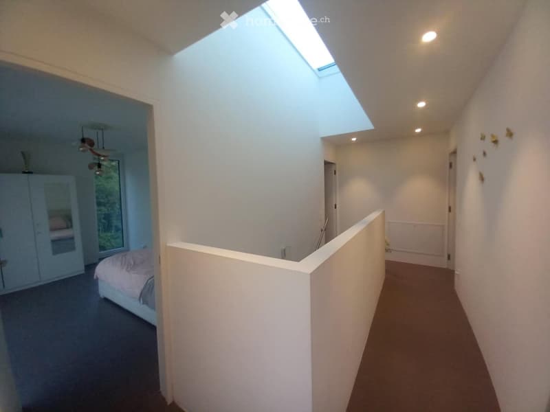 Bright hallway with a skylight ceiling, white walls, and carpeted floors. Features a raised section leading to a bedroom area with a bed and wardrobe visible in the background.