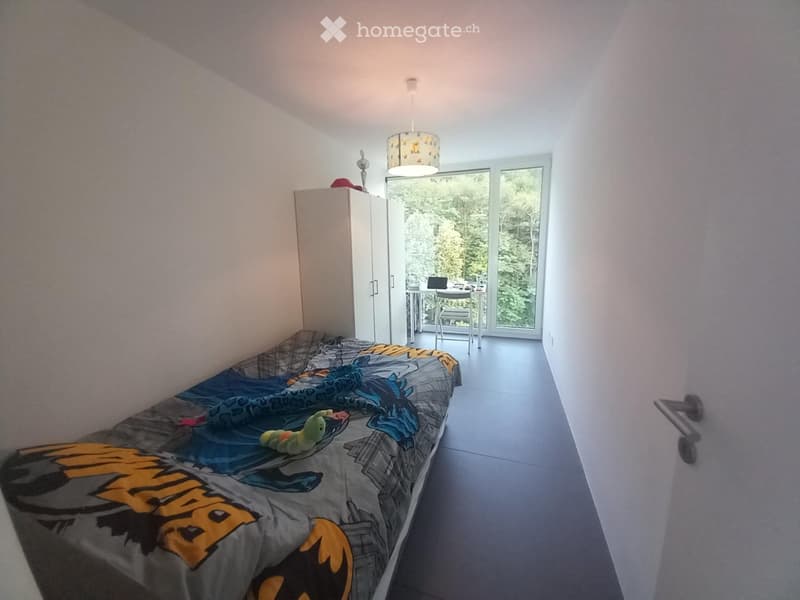 Bedroom with a single bed, colorful bedding, a wardrobe, a small table and chair near a large window with a view of greenery.