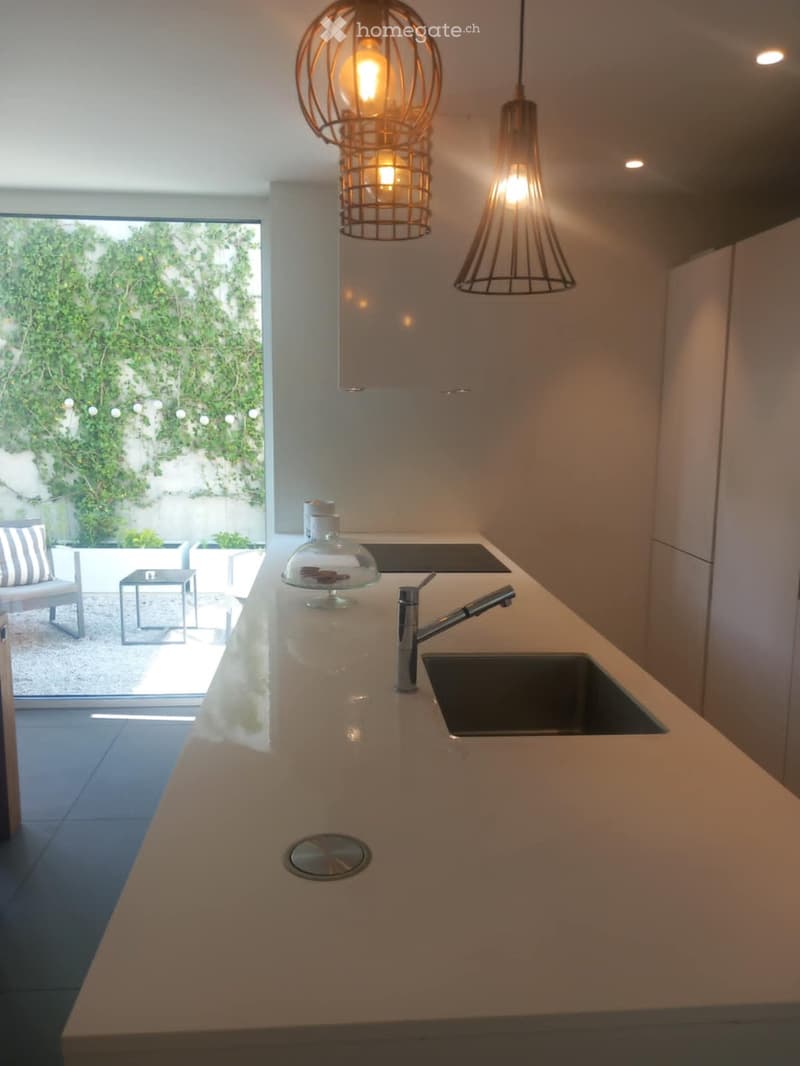 Modern kitchen with a light-colored countertop, stainless steel sink and faucet, and two hanging pendant lights. A glass bowl is placed on the counter, and the background shows a view of a garden through large windows.