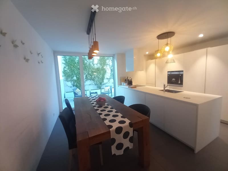 Modern kitchen with white cabinets, a central island, and a dining area with a wooden table and chairs. The space features pendant lighting and large windows overlooking an outdoor area.