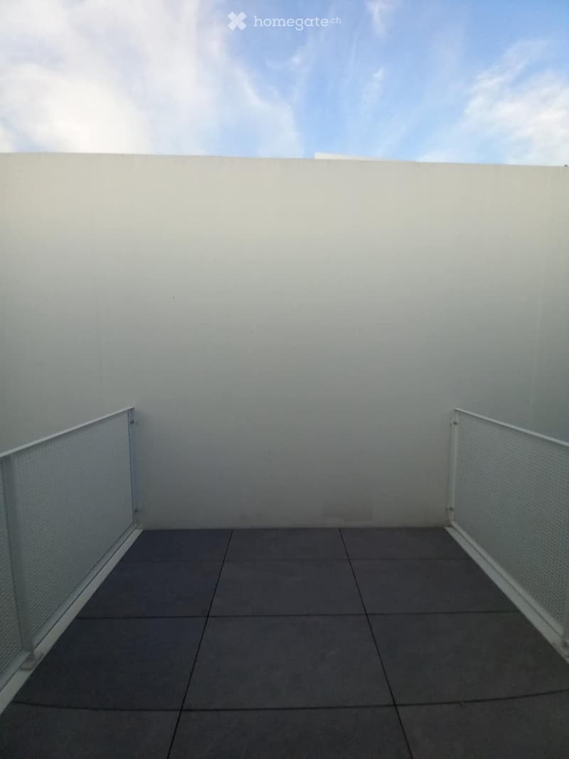 Empty balcony with grey tiled floor, white walls, and metal railings.