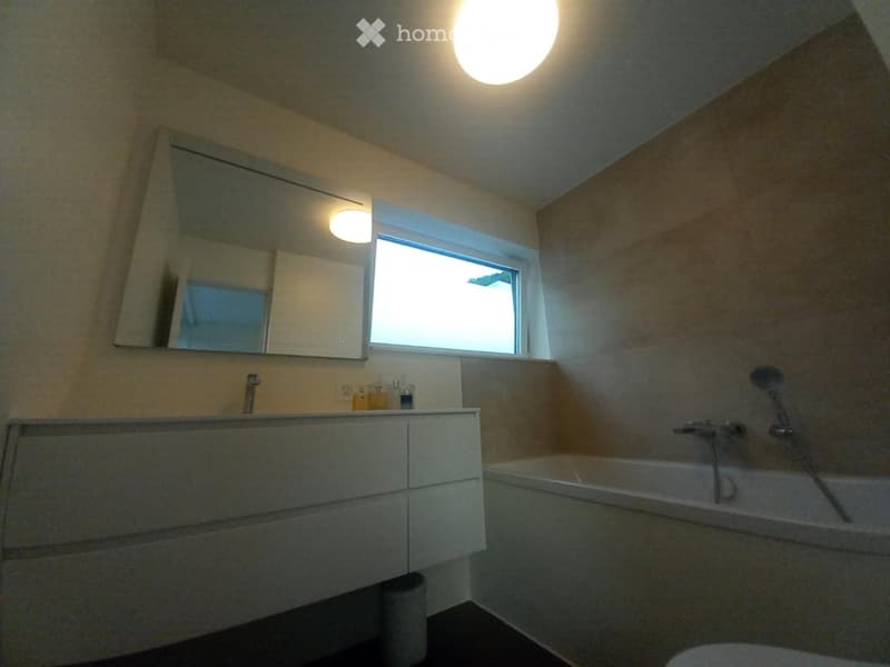 Modern bathroom with a large mirror, sink, bathtub, and showerhead. Features a window and built-in storage cabinet.