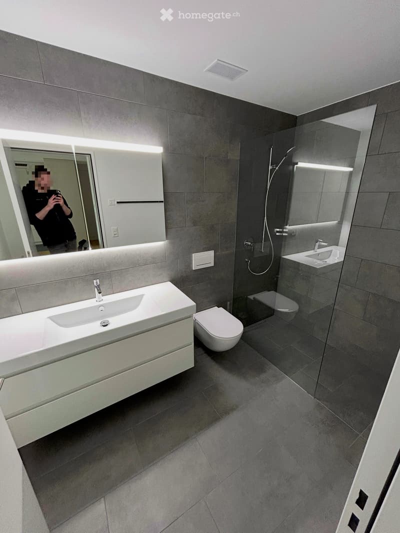 Modern bathroom with grey tiles, white sink, toilet, and large mirror with built-in lighting.