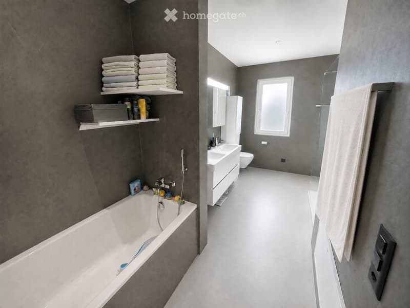 Modern bathroom with dark grey walls, white bathtub, sink, toilet, and shelves holding towels and toiletries.