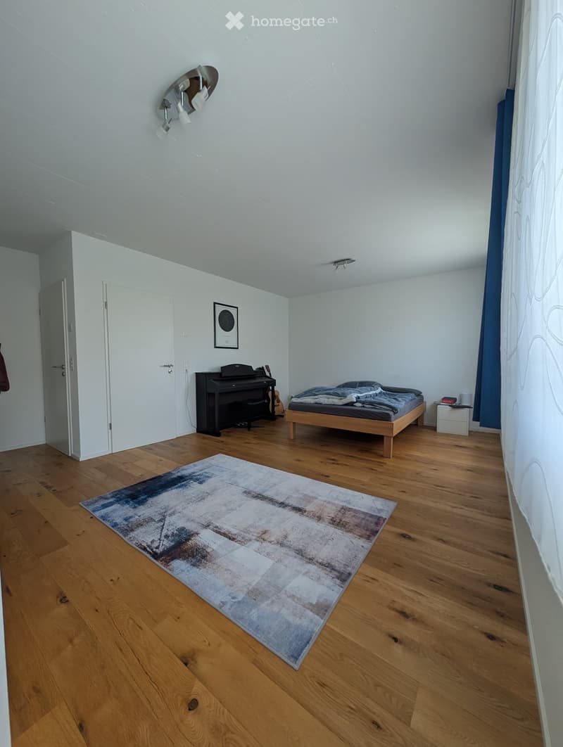 Bright bedroom with wooden flooring, a bed with blue bedding, a piano, a guitar, a nightstand, and a large abstract rug.