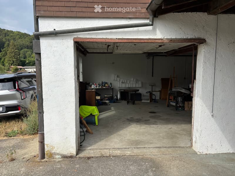 Garage with a concrete floor, white walls, and various stored items including a table, shelves, and a covered object.