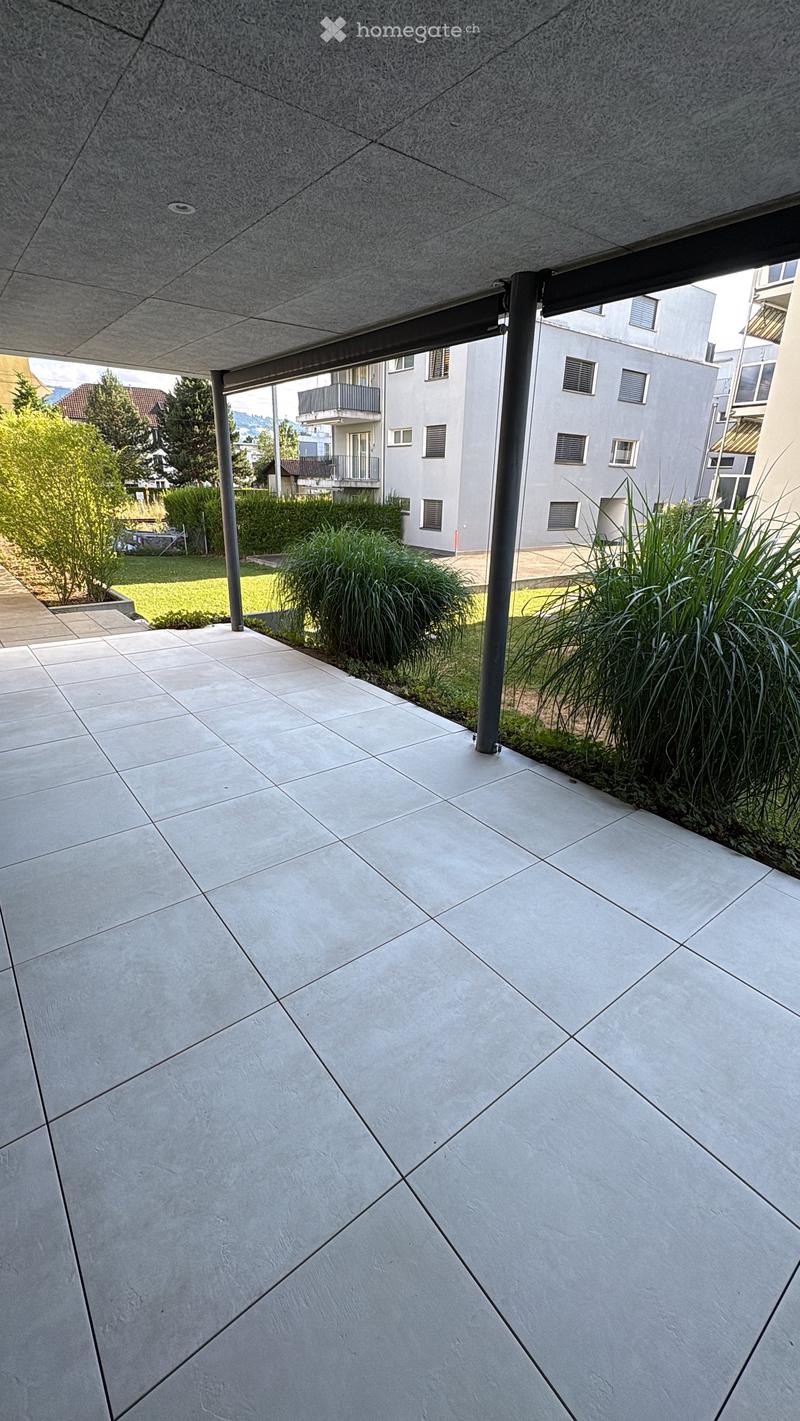 Modern outdoor patio with large, light-colored tiles and a covered area supported by metal poles. The space is bordered by greenery and leads to a grassy area with additional plants and trees.