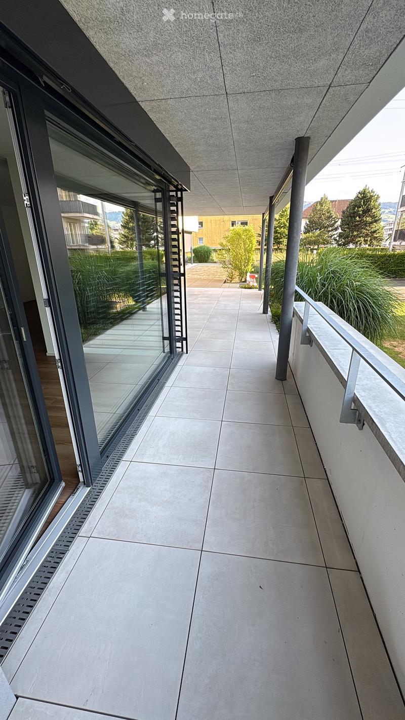 Modern balcony with tiled floor, glass railings, and a view of greenery and buildings.