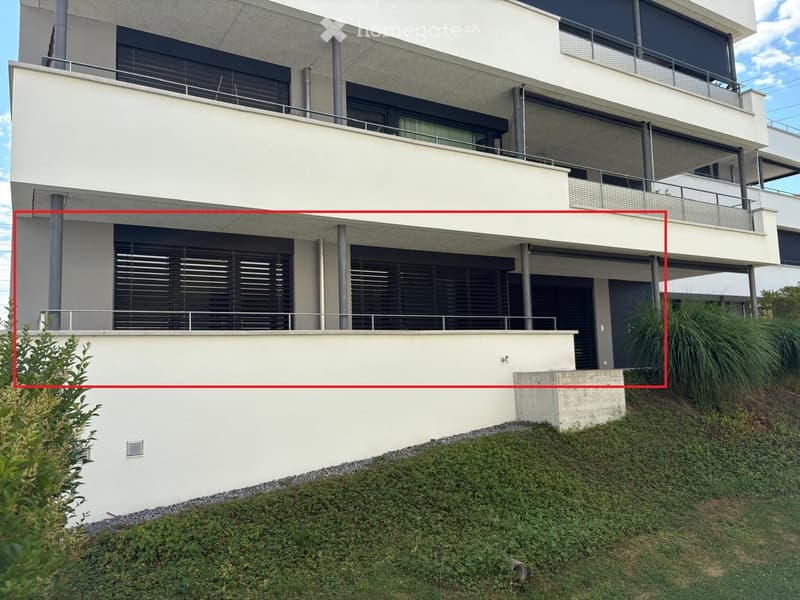 Modern two-story building with white facade, large windows, and balconies. The lower level has a raised ground floor with a concrete platform and greenery.