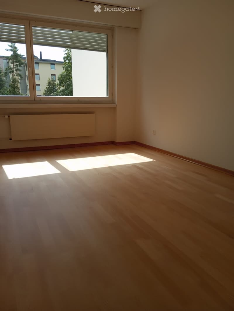 Empty room with wooden flooring, large window with blinds, radiator, and natural light streaming in.